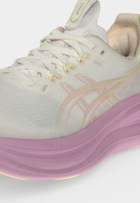 Κοντινή λήψη λευκού παπουτσιού τρεξίματος Asics με παστέλ ροζ σόλα, λογότυπο ανοιχτού πορτοκαλί και λευκά κορδόνια σε απλό φόντο.