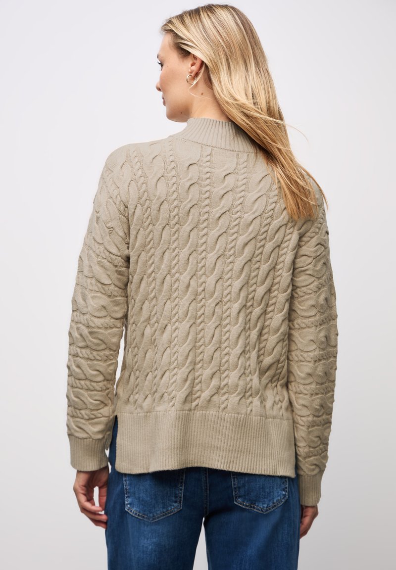 Beige Kabelstrickpullover mit strukturiertem Muster, geripptem Saum und Bündchen. Über blauen Jeans getragen, was eine lässige Passform zeigt.