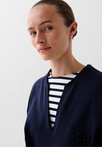 Marineblauwe pullover met V-hals, zachte textuur, voorzien van geborduurd "SCP" logo. Daaronder is een gestreept shirt in wit en marineblauw zichtbaar.