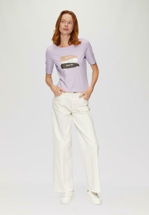 Femme aux cheveux roux portant une chemise lavande avec un texte et un pantalon large blanc, se tenant avec les mains dans les poches sur un fond uni.
