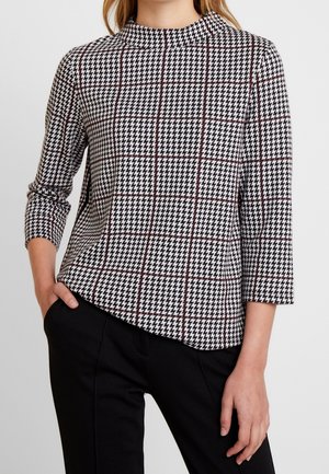 Femme portant un haut à motifs pied-de-poule noir et blanc avec des manches trois-quarts et un pantalon noir, la main dans la poche.