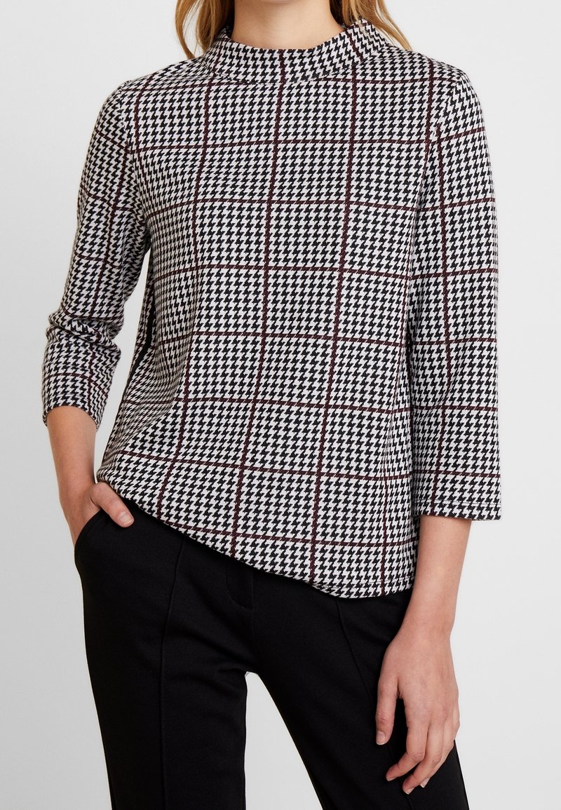 Donna che indossa una maglietta a motivo houndstooth bianco e nero con maniche a tre quarti e pantaloni neri, con una mano in tasca.