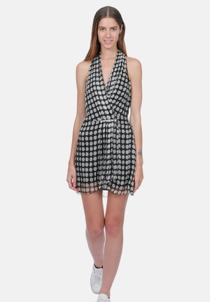 Femme portant une mini-robe noire sans manches à pois blancs et des baskets blanches, debout sur un fond uni.