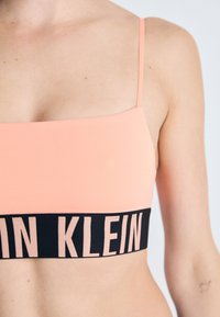 Perzik crop top met dunne bandjes, voorzien van een zwarte elastische band met het witte "CK" logo. Glad textuur en vierkante halslijn.