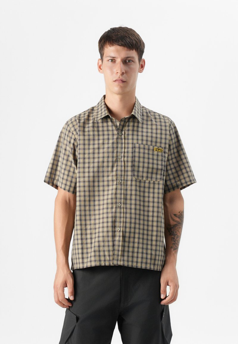 Young man stands wearing a short-sleeved plaid shirt with a pocket and logo, paired with dark pants, against a plain white background.