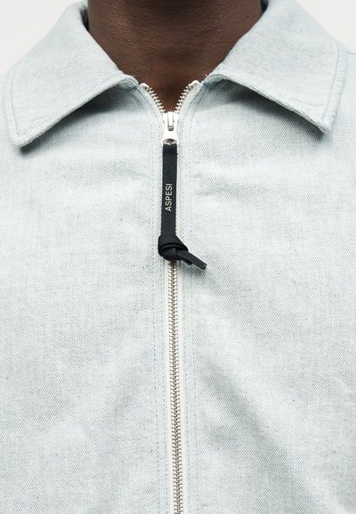 Ανοιχτό μπλε τζιν μπουφάν με ασημένιο φερμουάρ και μαύρη υφασμάτινη ετικέτα με την ένδειξη «ASPES» που φορά άτομο με σκούρο δέρμα.