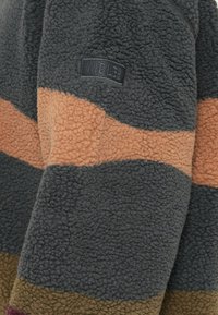 Fleecejacke mit auffälligen horizontalen Streifen in Grau, Rost und Brauntönen; strukturierter Stoff mit einem Patch-Logo.