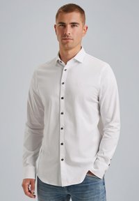 Weißes Langarmhemd mit Knöpfen aus Baumwolle, ausgestattet mit schwarzen Knöpfen und einem dezenten Logo auf der Brust, kombiniert mit blauen Jeans.