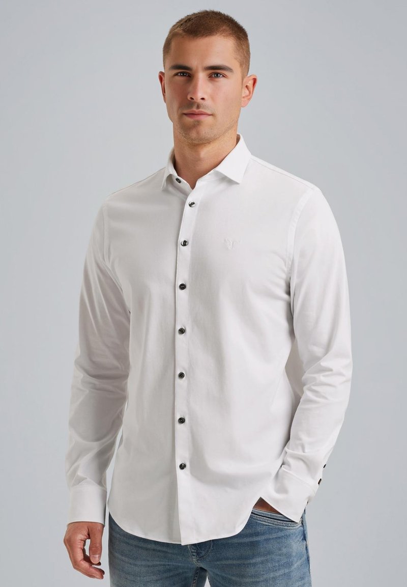 Weißes Langarmhemd mit Knöpfen aus Baumwolle, ausgestattet mit schwarzen Knöpfen und einem dezenten Logo auf der Brust, kombiniert mit blauen Jeans.