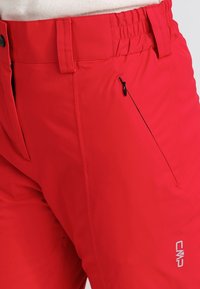Pantalon d'extérieur rouge avec une taille élastique, une poche latérale zippée et un logo de marque. Le tissu semble lisse et léger.