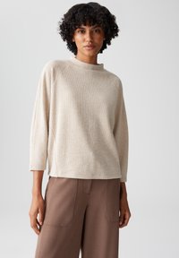 Beige, textured trui met een hoge hals en driekwart mouwen, gecombineerd met bruine, op maat gemaakte broeken met opgestikte zakken.