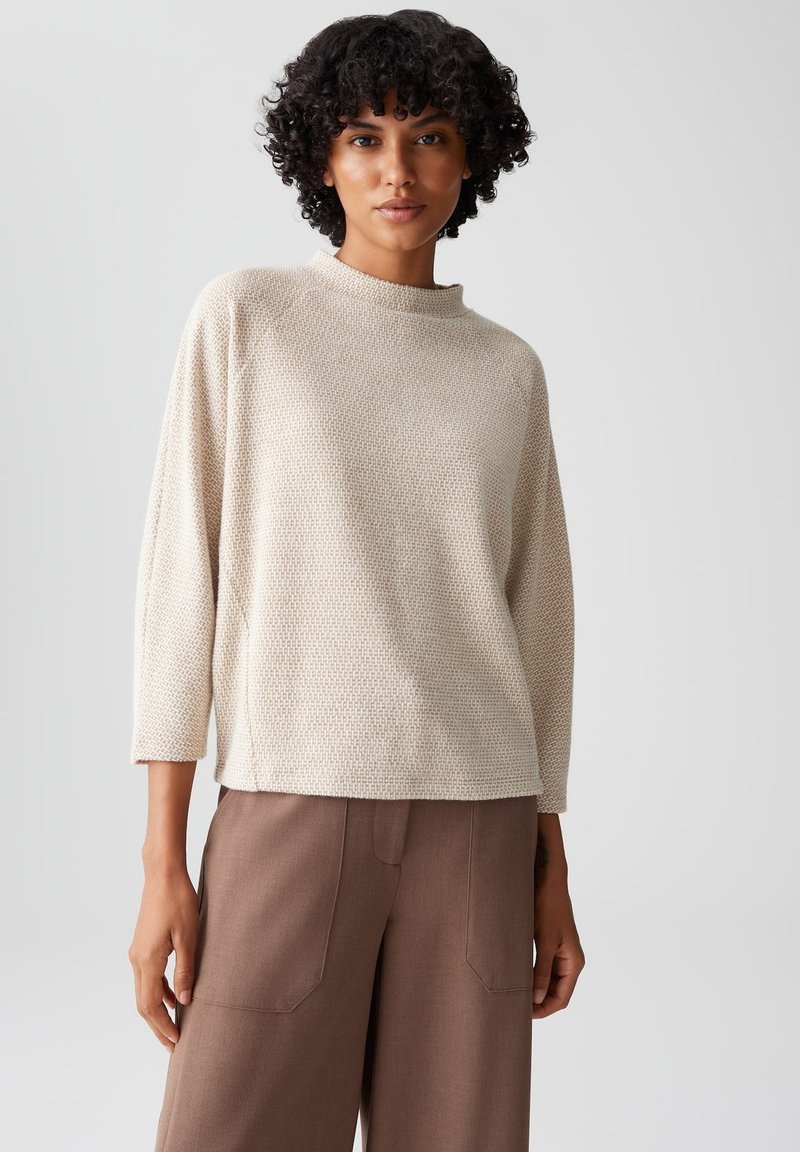 Beige, textured trui met een hoge hals en driekwart mouwen, gecombineerd met bruine, op maat gemaakte broeken met opgestikte zakken.