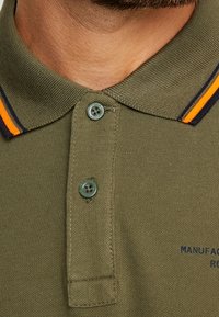 Koszulka polo w oliwkowym kolorze z bawełny o fakturowanej tkaninie. Posiada dwuknopowy zamek, kołnierzyk w niebiesko-pomarańczowe paski oraz haftowane logo.