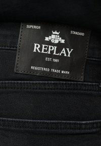 Μαύρο τζιν με ορθογώνιο μαύρο δερμάτινο λογότυπο που αναγράφει "REPLAY", "EST. 1981", "SUPERIOR", "STANDARD" και "REGISTERED TRADE MARK".