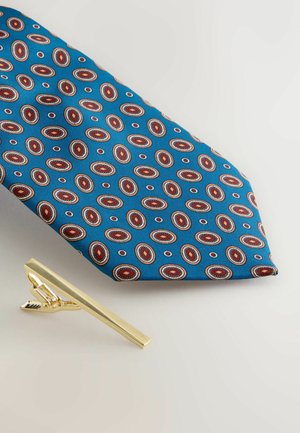 Blue tie with oval patterns in red and white; accompanied by a shiny gold tie clip with a rectangular shape and a clasp mechanism.