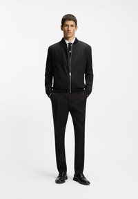 Schwarze Bomberjacke mit gerippten Bündchen und Kragen, silbernem Reißverschluss, getragen über einem weißen Hemd und schwarzen Hosen, kombiniert mit schwarzen Schuhen.