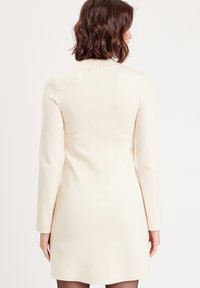 Robe à manches longues de couleur crème en tissu doux, présentant une silhouette cintrée et un design minimaliste, vue de dos.
