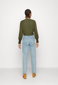 Maglione verde oliva con maniche a palloncino, abbinato a jeans chiari a gamba stretta. Scarpe marroni con tacco blocco, tasche posteriori visibili sui jeans.