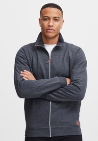 Sudadera de cremallera gris oscuro con cuello alto, puños y dobladillo acanalados. Presenta una etiqueta de marca discreta en la cadera izquierda. Textura de tela suave.