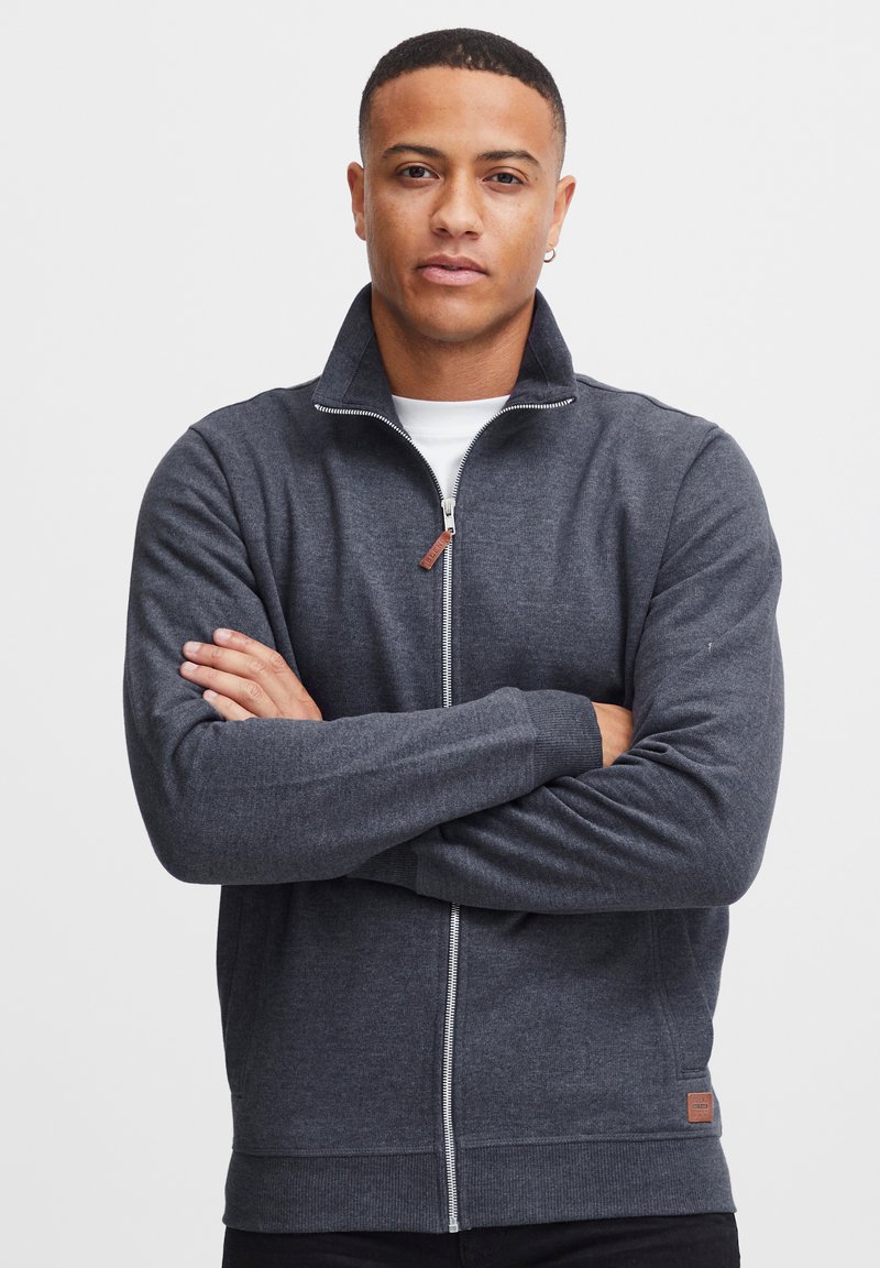 Sudadera de cremallera gris oscuro con cuello alto, puños y dobladillo acanalados. Presenta una etiqueta de marca discreta en la cadera izquierda. Textura de tela suave.