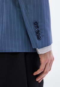 Sacou bleu deschis cu dungi subțiri, cu o textură netedă, prevăzut cu patru nasturi închiși la culoare pe manșetă și o manșetă de cămașă albă vizibilă dedesubt.