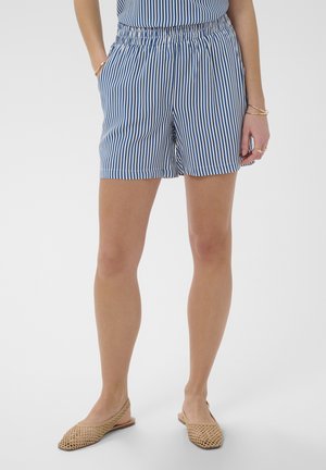Jambes portant un short rayé vertical bleu et blanc et des chaussures slingback tissées couleur beige, les mains reposant le long du corps sur un fond uni.