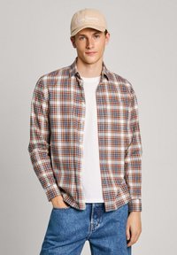 Camisa a cuadros en tonos de beige, marrón y azul; diseño de botones; sobre una camiseta blanca; combinada con vaqueros de un azul claro y una gorra beige.