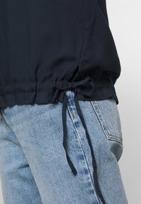 Nahaufnahme von hellblauen Denim-Jeans mit einem dunkelblauen Oberteil, das an der Taille mit einem gebundenen Kordelzug versehen ist, vor einem schlichten Hintergrund.