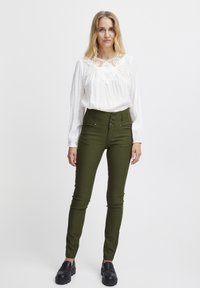 Blusa blanca con detalles de encaje en el cuello, combinada con pantalones verde oliva de talle alto y zapatos negros de plataforma.