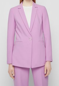 Levandulový blazer s klopami, dvěma předními kapsami, zapínáním na jeden knoflík a detaily ze stříbrného kovu, spárovaný s bílou vnitřní blůzou.