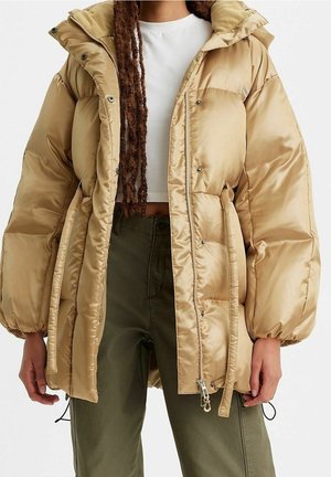 Osoba ubrana w beżową puchową kurtkę na białej krótkiej bluzce i oliwkowozielonych spodniach, stojąca na jednolitym tle.