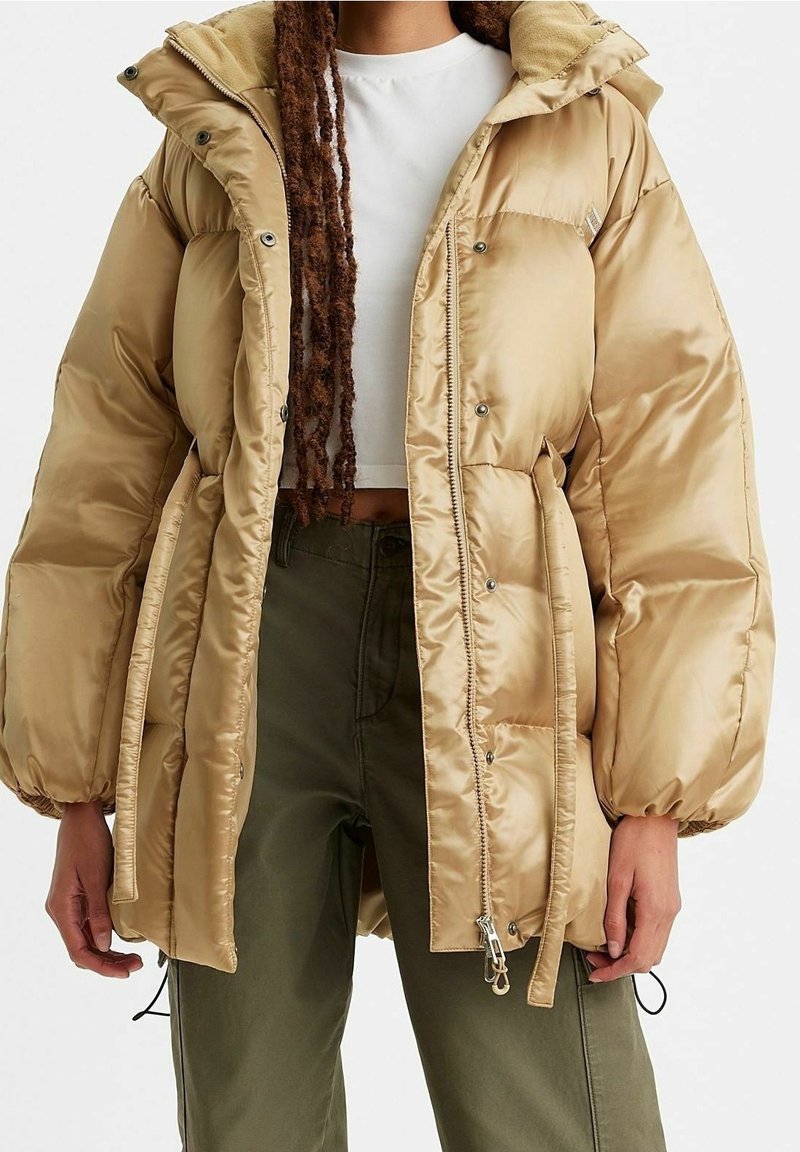 Osoba ubrana w beżową puchową kurtkę na białej krótkiej bluzce i oliwkowozielonych spodniach, stojąca na jednolitym tle.