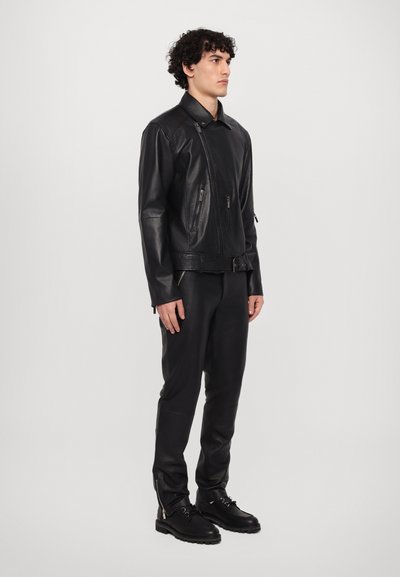 Zwarte leren jas met een rits, zijzakken en een riemdetail. Gecombineerd met strakke zwarte broek en zwarte veterschoenen.