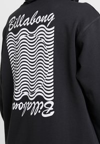 Sudadera negra con un gráfico de línea ondulada blanca en negrita y el texto "Billabong", con un acabado texturizado y un corte relajado.