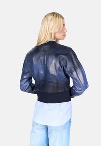 Zkrácená modrá kožená bomber bunda s žebrovaným námořnickým lemem, se subtilním leskem a logem na zadní straně, nošená přes pruhovanou košili.