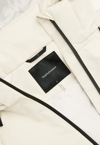 Lys beige jakke med sort lynlås, åben for at afsløre sort Peak Performance label og størrelse S mærke indeni.
