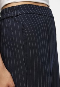 Marinblå randiga byxor med elastisk midja, utrustade med sidofickor och en slät, texturerad tyg. Rena linjer och subtil lyster.