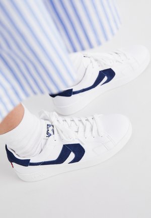 Pies con zapatillas blancas Levi's con detalles en azul marino y calcetines blancos, combinados con pantalones a rayas azules y blancas.