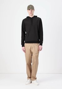 Hanorac negru cu buzunar frontal și șnururi, asociat cu pantaloni bej și adidași albi. Modelul poartă o pălărie verde. Look simplu, casual.