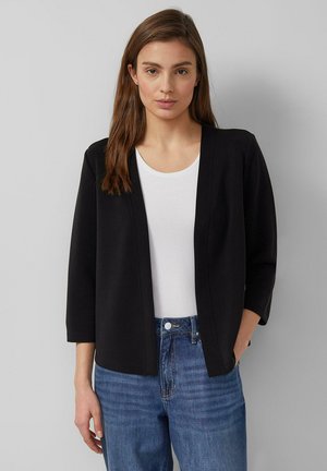 Vrouw draagt een zwart openvallend colbert, een wit shirt met een ronde hals en een hoge taille blauwe spijkerbroek, staand tegen een effen lichtgrijze achtergrond.