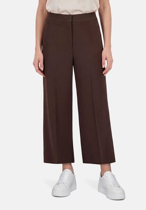 Femme portant un pantalon large marron foncé court et des baskets blanches à plateforme, debout sur un fond blanc.