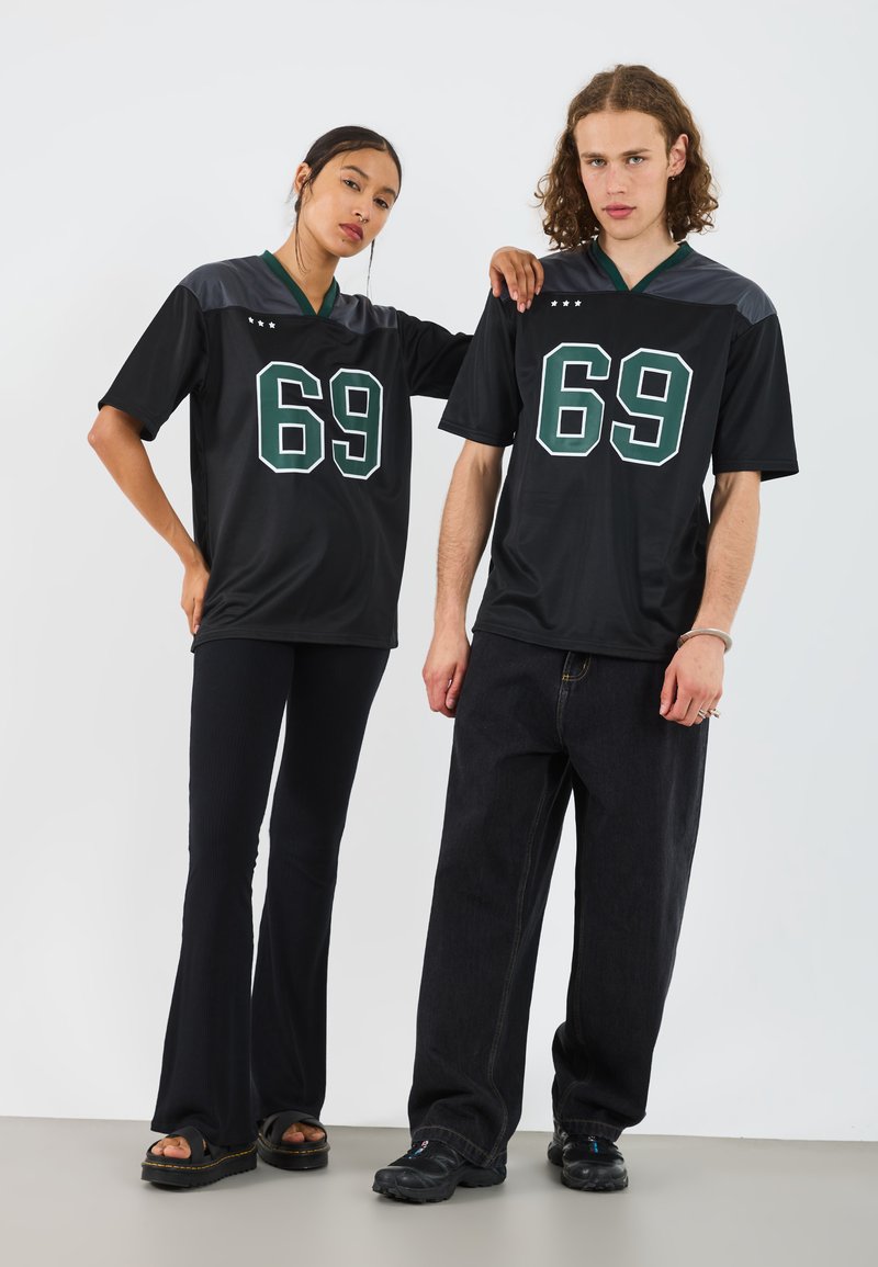 Camiseta negra de manga corta con detalles verdes y un gran "69" en el frente, combinada con pantalones de mezclilla anchos oscuros. El modelo posa junto a ella.