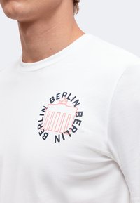 Gros plan d'une personne portant un t-shirt blanc avec un contour rose de la Porte de Brandebourg et le mot Berlin répété autour en lettres noires.