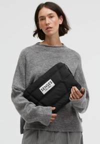 Schwarze, gesteppte Clutch mit einem Logolabel, gehalten von einer Person in einem grauen Pullover, die eine weiche Textur und rechteckige Form zeigt.