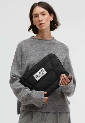 Black quilted clutch with a logo label, held by a person in a grey sweater, showcasing a soft texture and rectangular shape.