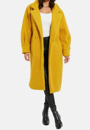 Kvinde iført en lang, sennepsgul, tekstureret frakke over en hvid top, sorte læderbukser og sorte ankelstøvler, stående med benene krydsede.