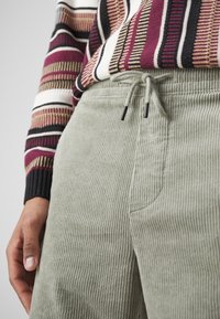 Green corduroy pants with a textured finish, featuring an elastic waistband and black drawstring, paired with a multicolored striped sweater.