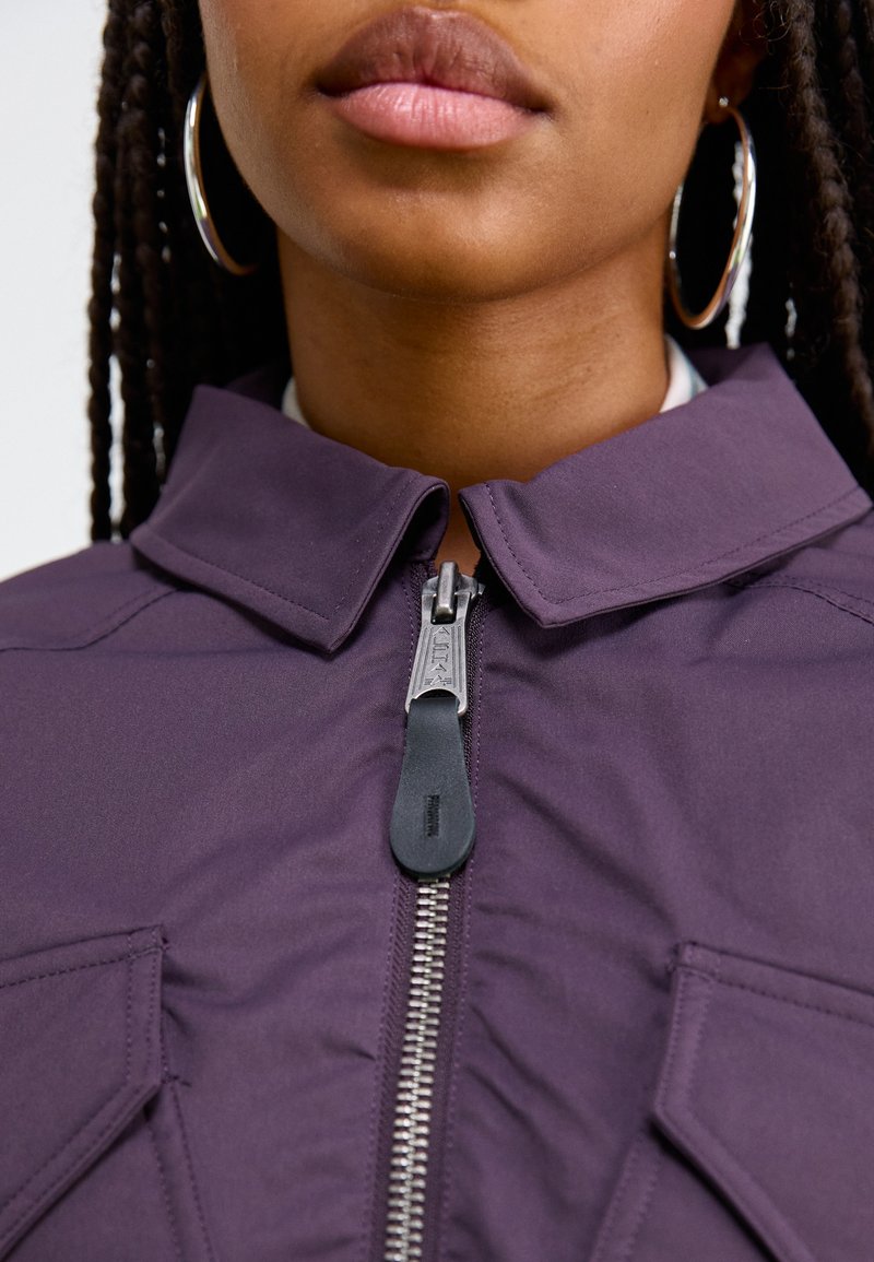 Chaqueta de cremallera morada con cuello tipo camisa, que presenta una cremallera plateada y bolsillos laterales. Tejido suave con una textura blanda.