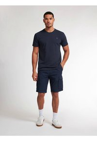 Vīrietis valkā tumši zilu T-kreklu, tumši zilas kargo šortus, baltas zeķes un baltas sporta kurpes, stāvot pret vienkāršu gaišu fonu.