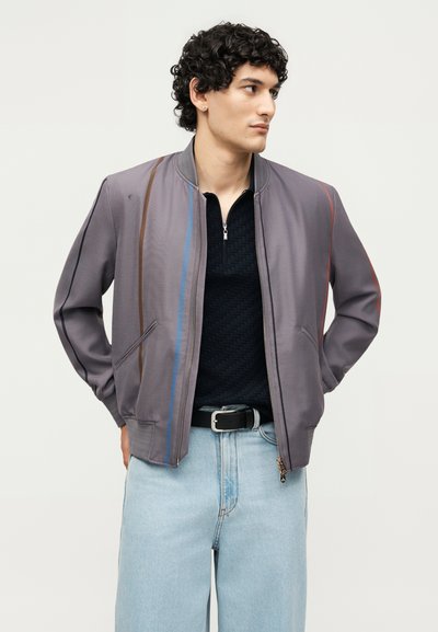 Νέος άντρας που φοράει γκρι σακάκι με κάθετες πολύχρωμες ρίγες, μαύρο υφασμάτινο πουκάμισο, ανοιχτό μπλε τζιν και μαύρη ζώνη, κοιτάζει προς τα πλάγια.